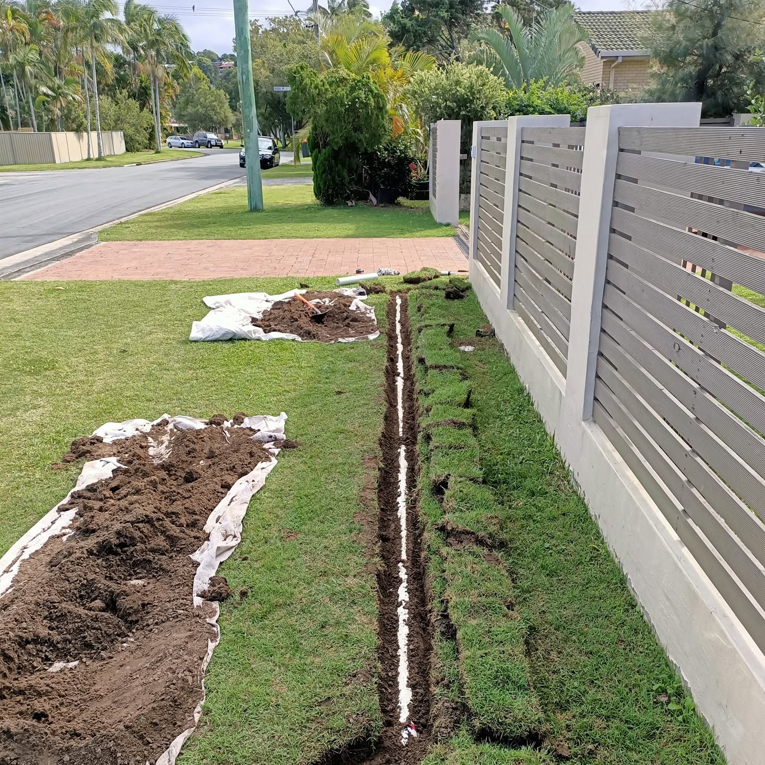 A trench dug along a front lawn and fence line for new pipework, turf rolled back